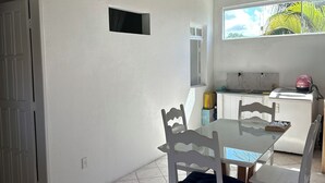 Un comedor con una mesa y sillas blancas, piso de baldosas, ventana con vistas, y una zona de cocina al fondo.