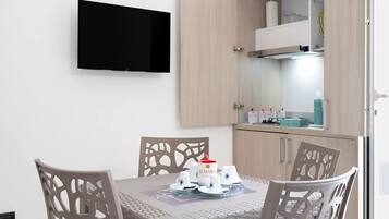 A dining area with a table set for four, a flat-screen TV mounted on the wall, and light-colored wooden cabinets with a kitchen area visible in the background.