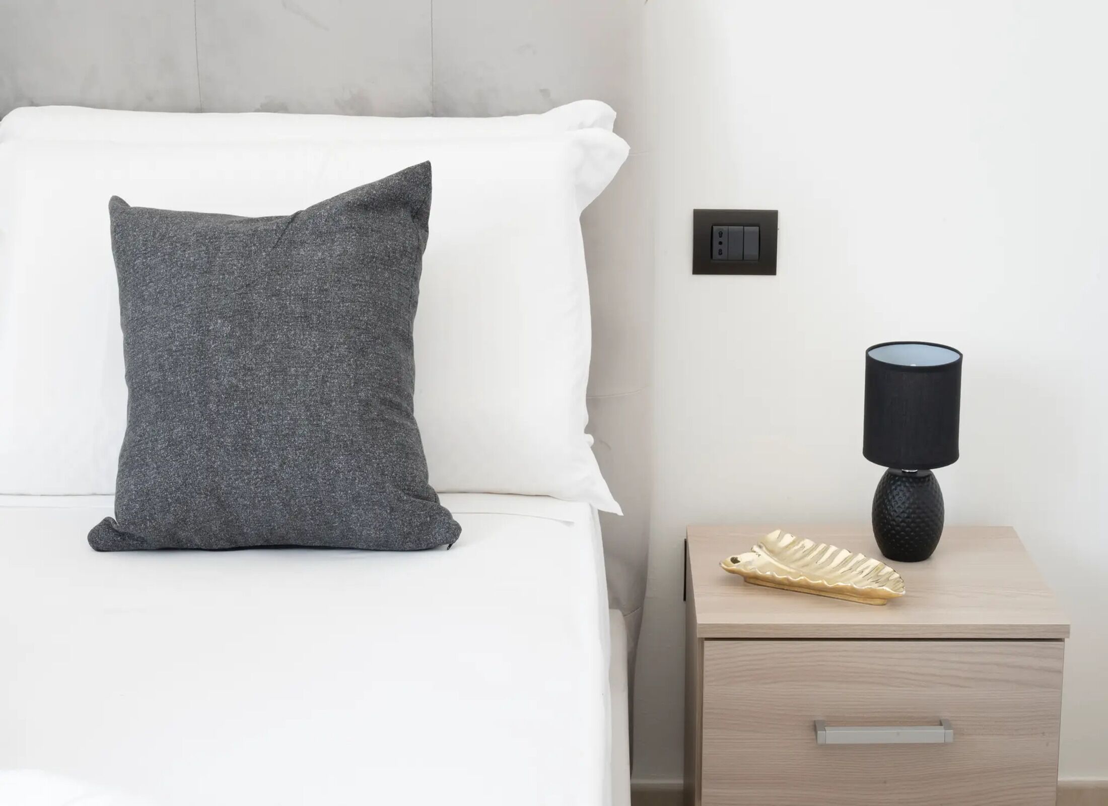 A bed with a grey pillow, a bedside lamp, and a small drawer unit.