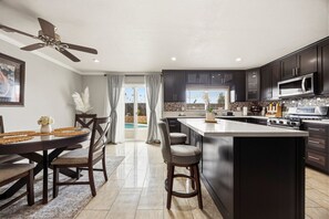 A modern kitchen with dark cabinetry, a central island, a dining area with a table and chairs, and a ceiling fan. - Charming 3BR w/ Hills Views, Pool & Billiards (Hemet)