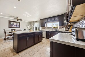 A modern kitchen with dark cabinetry, a large island, a dining area with chairs, and a ceiling fan. - Charming 3BR w/ Hills Views, Pool & Billiards (Hemet)