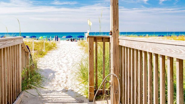 A wooden boardwalk leading to a beach with umbrellas and a clear sky. - Seacrest #507 (Gulf Shores)