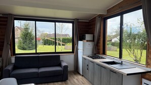A compact living space with a sofa, kitchenette, and large windows offering a view of a yard and trees. - Cocoon-style wooden accommodation with breakfasts included (Héloup)