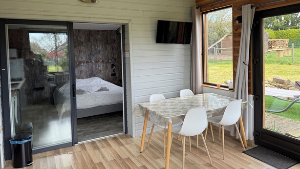 A modern room with a bed, a glass door leading to a balcony, a dining table with chairs, and a TV mounted on the wall. - Cocoon-style wooden accommodation with breakfasts included (Héloup)
