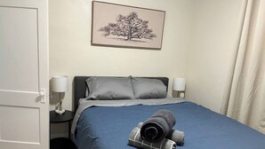 A bedroom with a bed, two bedside lamps, a door, a framed picture on the wall, and a rolled towel on the bed.