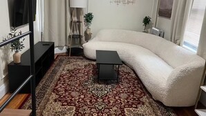 A modern living room with a large L-shaped sofa, a small black table, a patterned rug, a TV on a black stand, and a radiator.