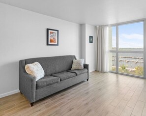 Un canapé gris avec deux coussins, un tableau encadré au mur et une grande fenêtre donnant sur un port de plaisance.