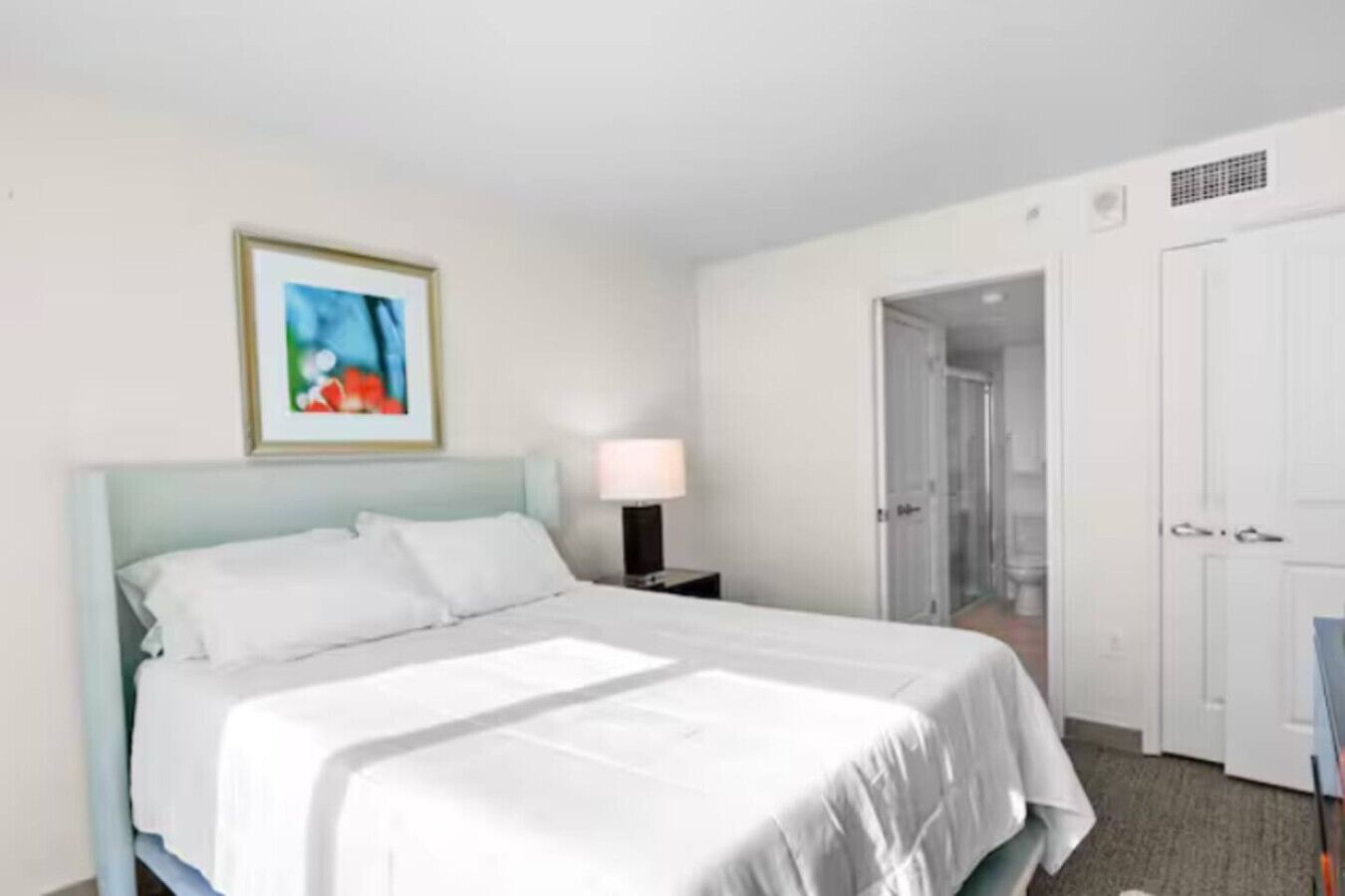 A hotel room with a bed, a nightstand with a lamp, a framed picture on the wall, and a door leading to a bathroom.