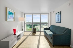 A modern living room with a sofa, a red vase, a lamp, and a large window offering a city view. - Modern Apt w/ City Views in Downtown Fort Myers (Fort Myers)