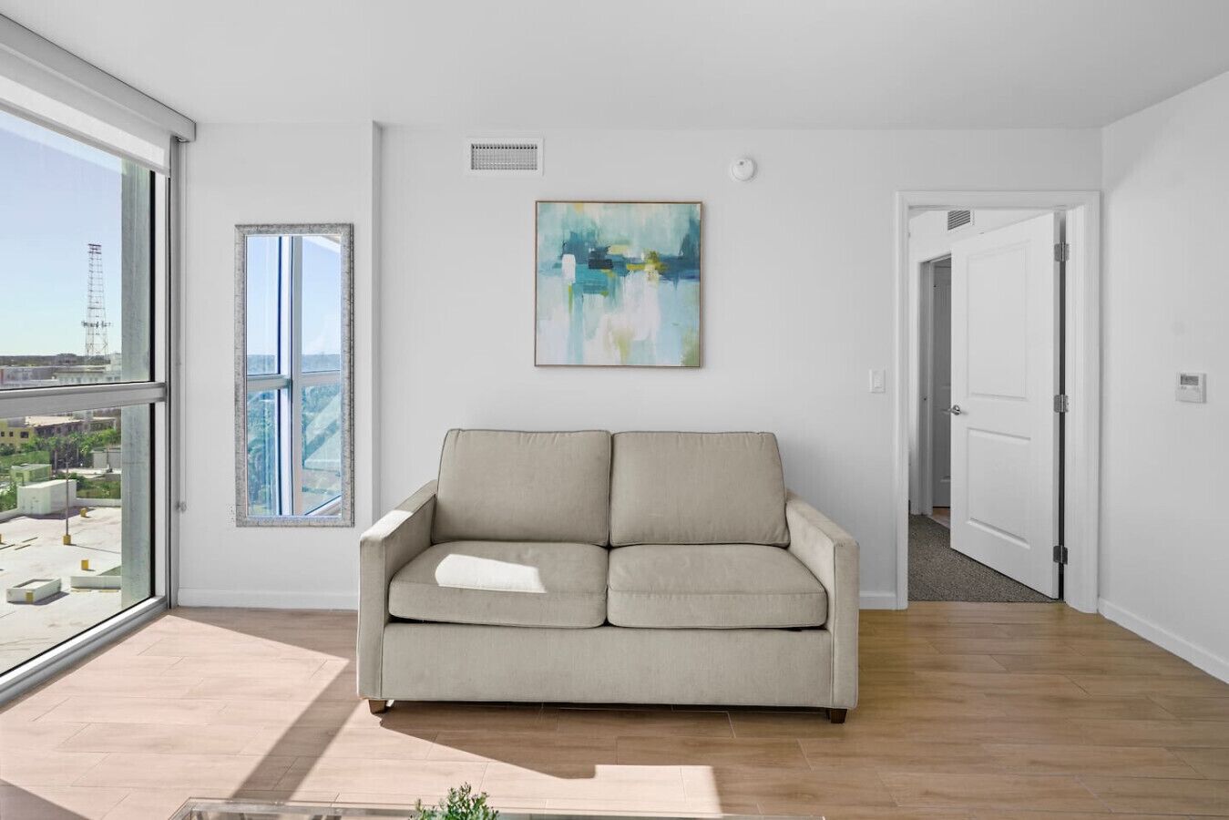 Una sala moderna con un sofá, un ventanal grande, un cuadro y una puerta que da a otra habitación.