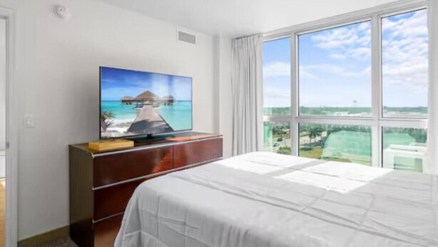 Ein Hotelzimmer mit einem Bett, einem Fernseher auf einer Holzkombination und einem großen Fenster mit Blick auf das Meer und Gebäude.