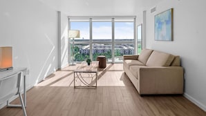 Ein modernes Wohnzimmer mit einer Couch, einem Couchtisch und einer Lampe. Ein großes Fenster bietet Blick auf Gebäude und Grünflächen.
