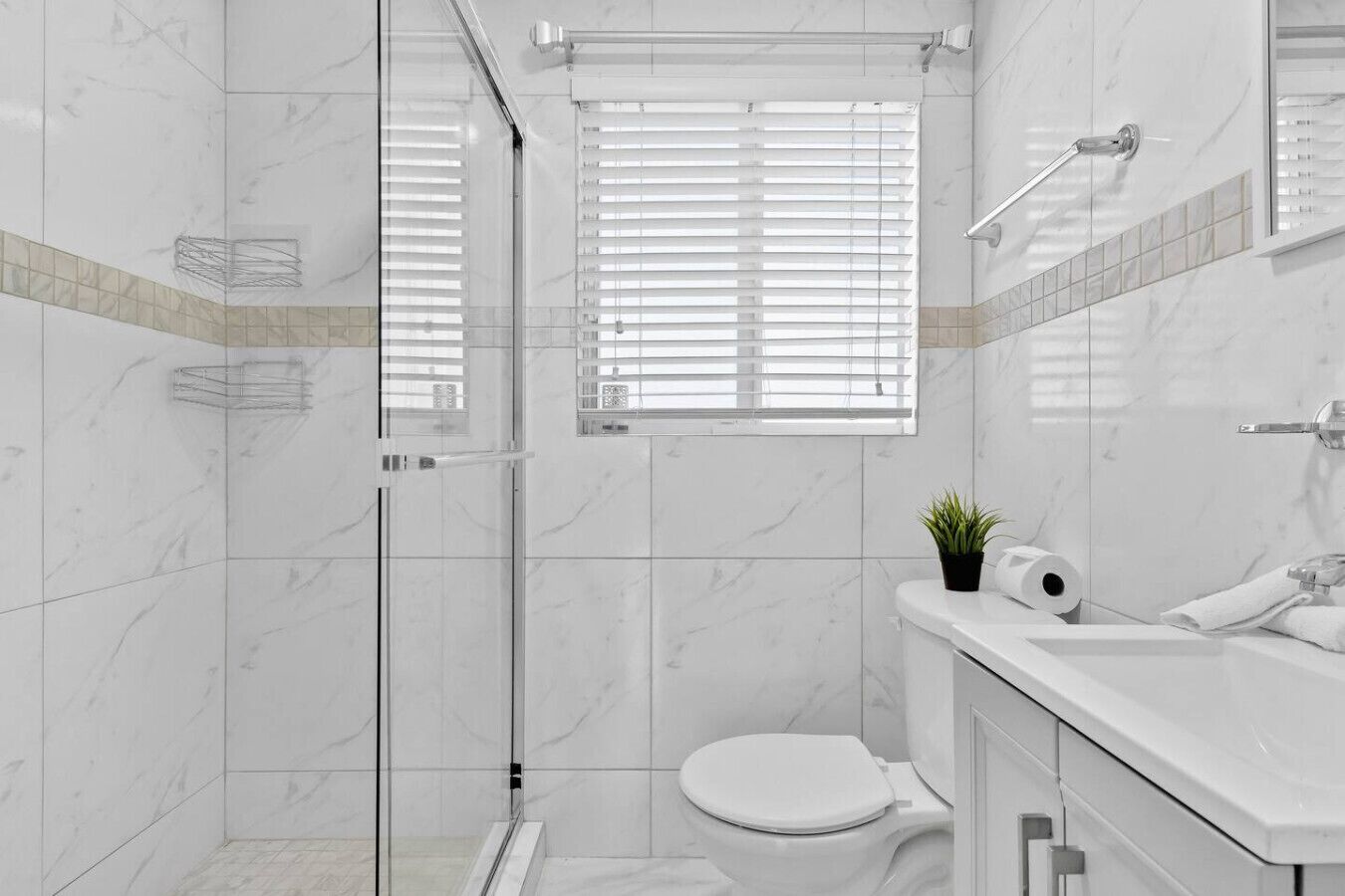 A modern bathroom with a glass shower enclosure, a toilet, a sink with a mirror, and a window with blinds.