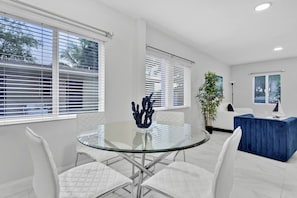 A modern living room with a glass table, white chairs, a blue sofa, and a potted plant. - Cozy Caribbean Retreat Near Wynwood & Design (Miami)