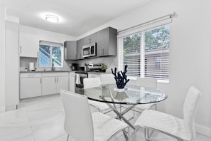 A modern kitchen with white cabinetry, a glass dining table, and a view through the window. - Cozy Caribbean Retreat Near Wynwood & Design (Miami)