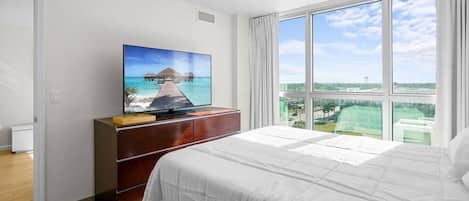 A hotel room with a bed, a TV on a wooden dresser, and a large window with a view of a beach and buildings.
