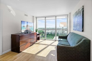Ein modernes Wohnzimmer mit großem Fenster, Flachbildfernseher, dunklem Holzschrank, grünem Sofa und einem gerahmten Bild an der Wand.