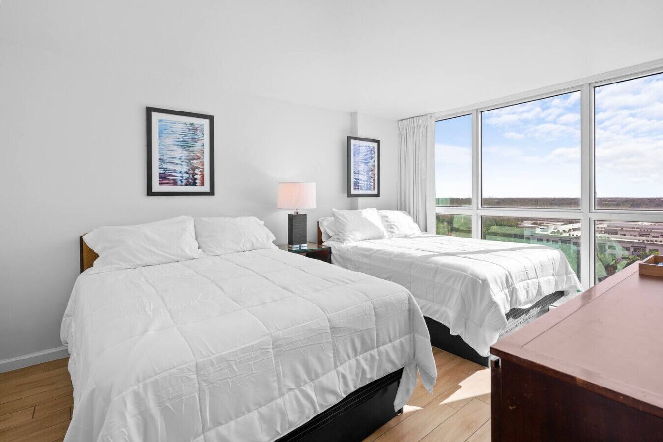 A hotel room with two beds, a desk, a window with a city view, and artwork on the walls.