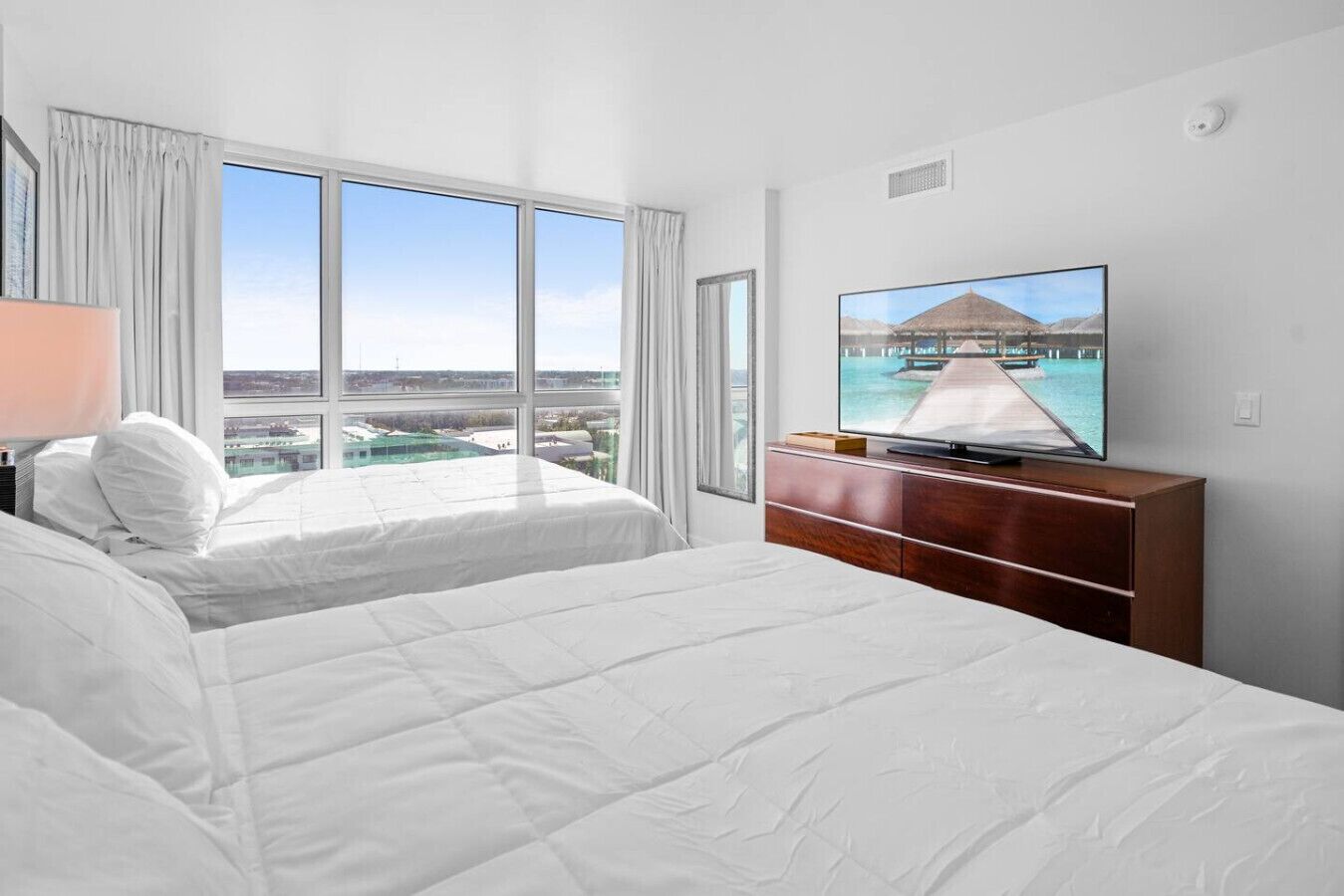 Une chambre d’hôtel avec deux lits, un téléviseur posé sur une commode en bois et une grande fenêtre donnant sur une jetée et la mer.