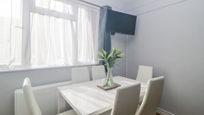 A dining area with a white table and chairs, a potted plant, and a window with sheer curtains. - Devon Seaside Apartment (Paignton)