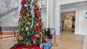 Een grote kerstboom in een lichte hal, versierd met decoraties en lichtjes, omringd door cadeaudozen, met een trap en moderne verlichtingsarmaturen.