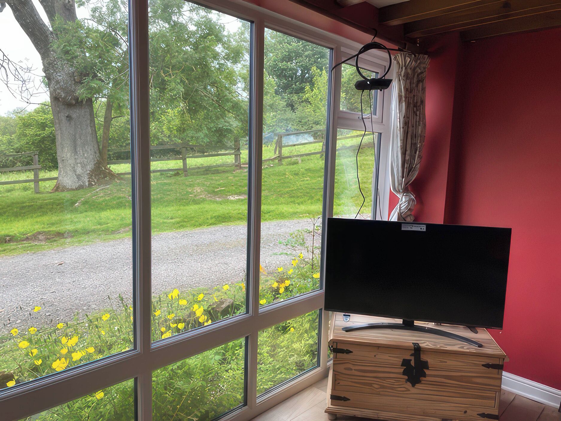 Ein Zimmer mit Fernseher auf einer Holzkommode, große Fenster mit Blick auf eine Rasenfläche und Bäume.