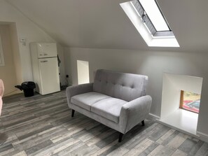 A room with a grey sofa, wooden flooring, a skylight, a white refrigerator, and a door. - The Cottage (Middlesbrough)