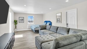 A spacious living room with a large grey sectional sofa, a bed with blue bedding, a potted plant, and a view of the outside through a window. - Penthouse Suite Steps to the Beach | Jersey Shore Stay (Seaside Heights)