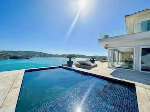 Una piscina con vistas al mar, una casa moderna con puertas de cristal y un cielo despejado.