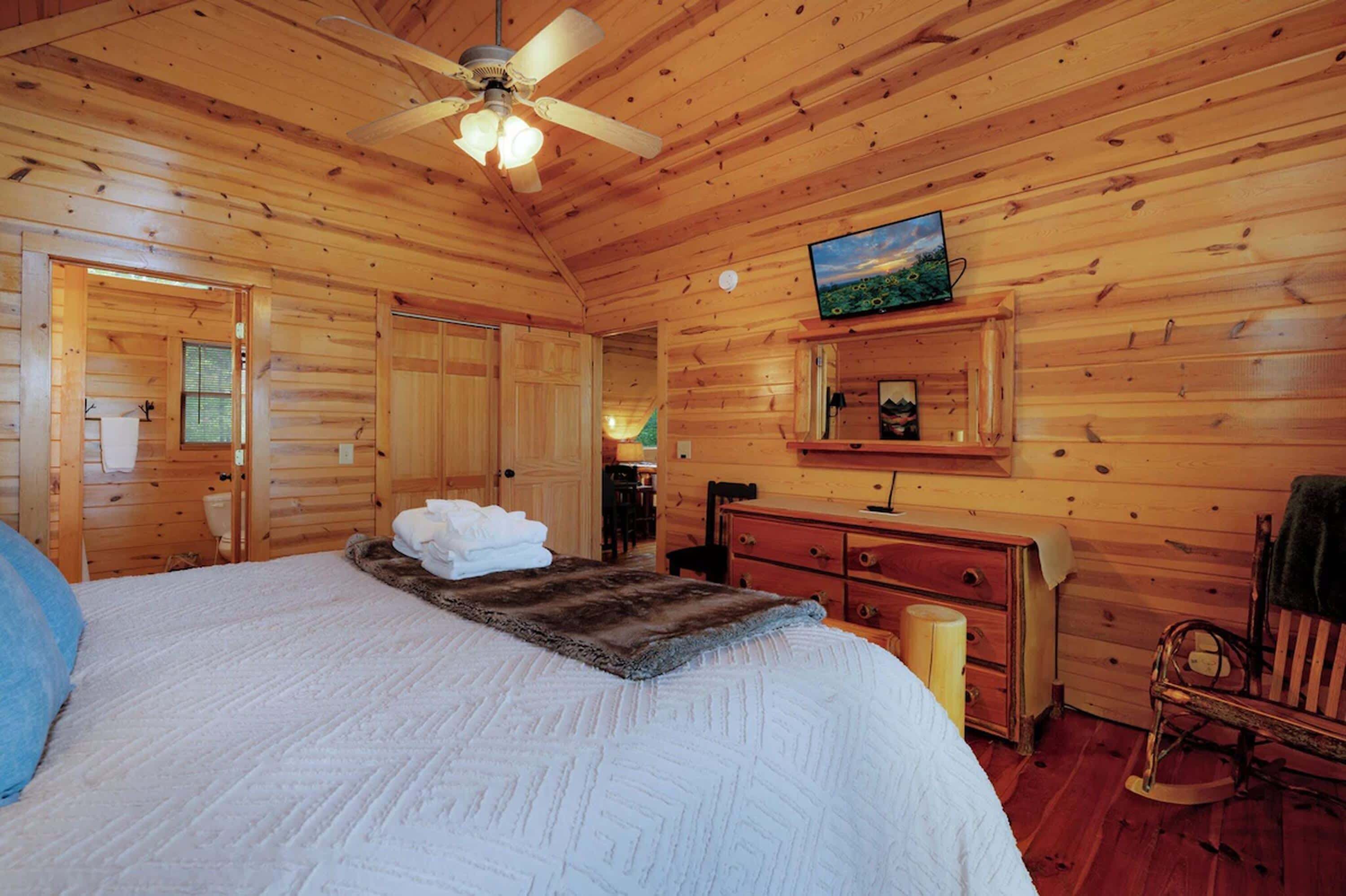 A wooden cabin room with a bed, a ceiling fan, a television, and a dresser.