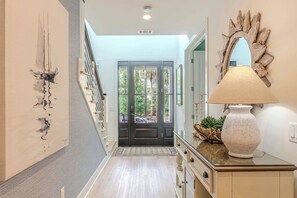 A hallway with a wooden floor, a staircase, a door with glass panels, a wall-mounted mirror, and a lamp on a cabinet. - Luxury Dunes Paradise - Private Pool (Hilton Head Island)