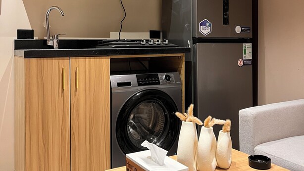 A compact kitchen with wooden cabinets, a microwave, a refrigerator, a washing machine, and a table with a tissue box and decorative items. - Perfect studio with AC in charming 6 October city (6 October)