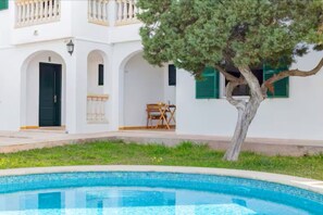 A white building with arched doorways, a tree in the foreground, and a swimming pool. - Nuria Cala Blanca (Cala Blanca)