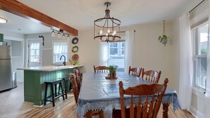 Un espace repas avec une grande table, des chaises en bois, un lustre et une cuisine en arrière-plan.