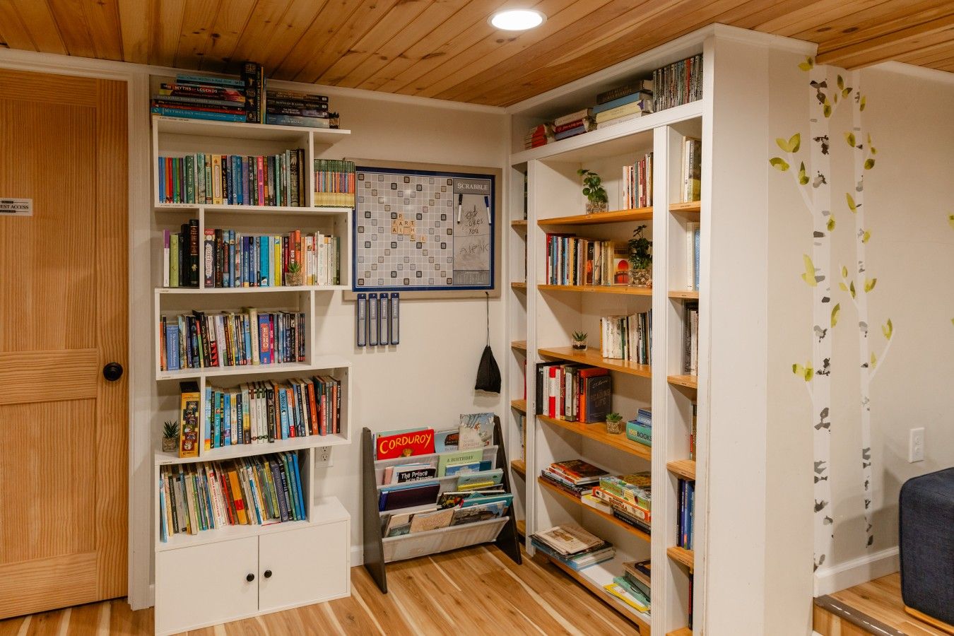 Habitación con suelo de madera, paredes blancas y múltiples estanterías llenas de libros. Hay un tablero de Scrabble en la pared y un cartel que indica 'First Access'.