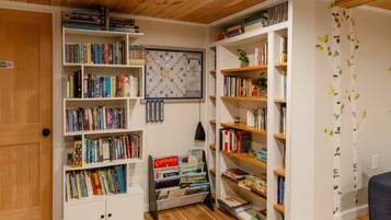 Habitación con suelo de madera, paredes blancas y múltiples estanterías llenas de libros. Hay un tablero de Scrabble en la pared y un cartel que indica 'First Access'.