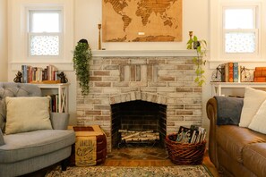 A cozy living room with a brick fireplace, a grey armchair, a brown leather sofa, and a bookshelf filled with books.