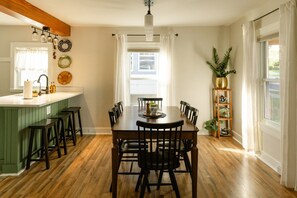 A dining area with a wooden table and chairs, a kitchen island with stools, and a window with curtains. - The Storybook Cottage | Close to Lancaster | Secret Room (Lancaster)