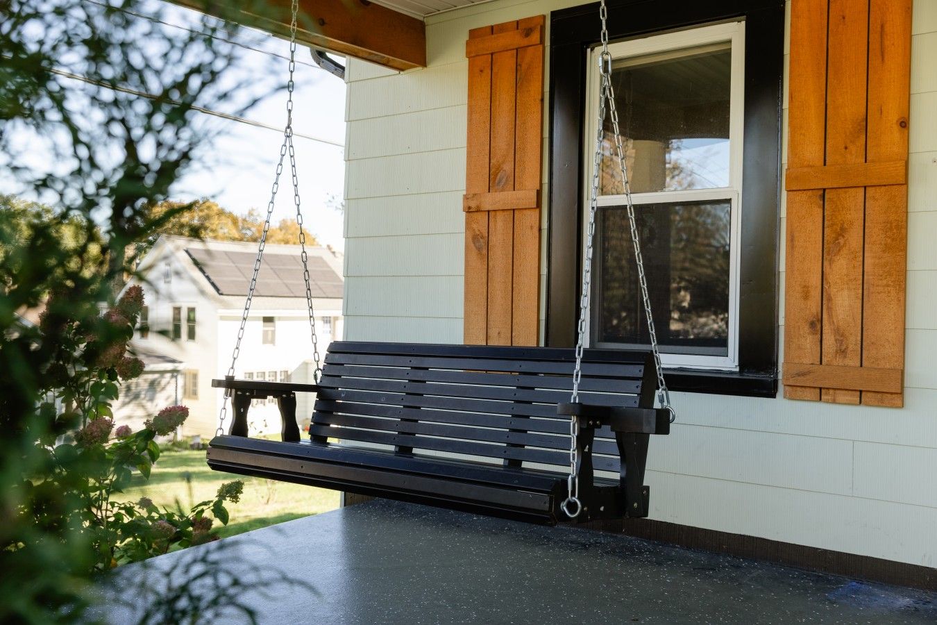 A porch with a swing bench, a window with shutters, and a view of a house in the background.