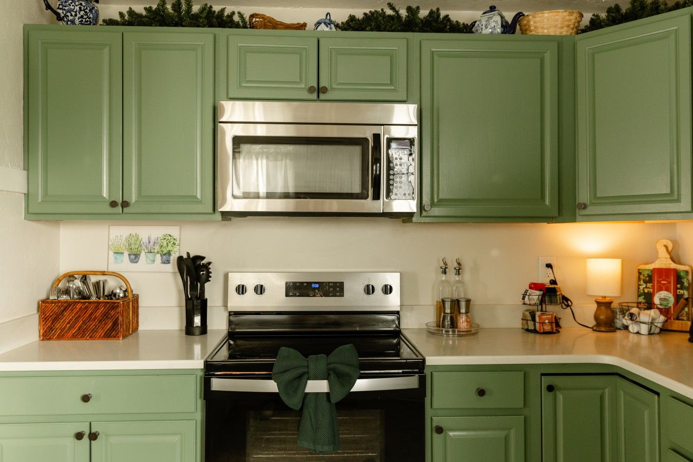 A kitchen with green cabinets, a stainless steel microwave, a black oven, and a wooden utensil basket.