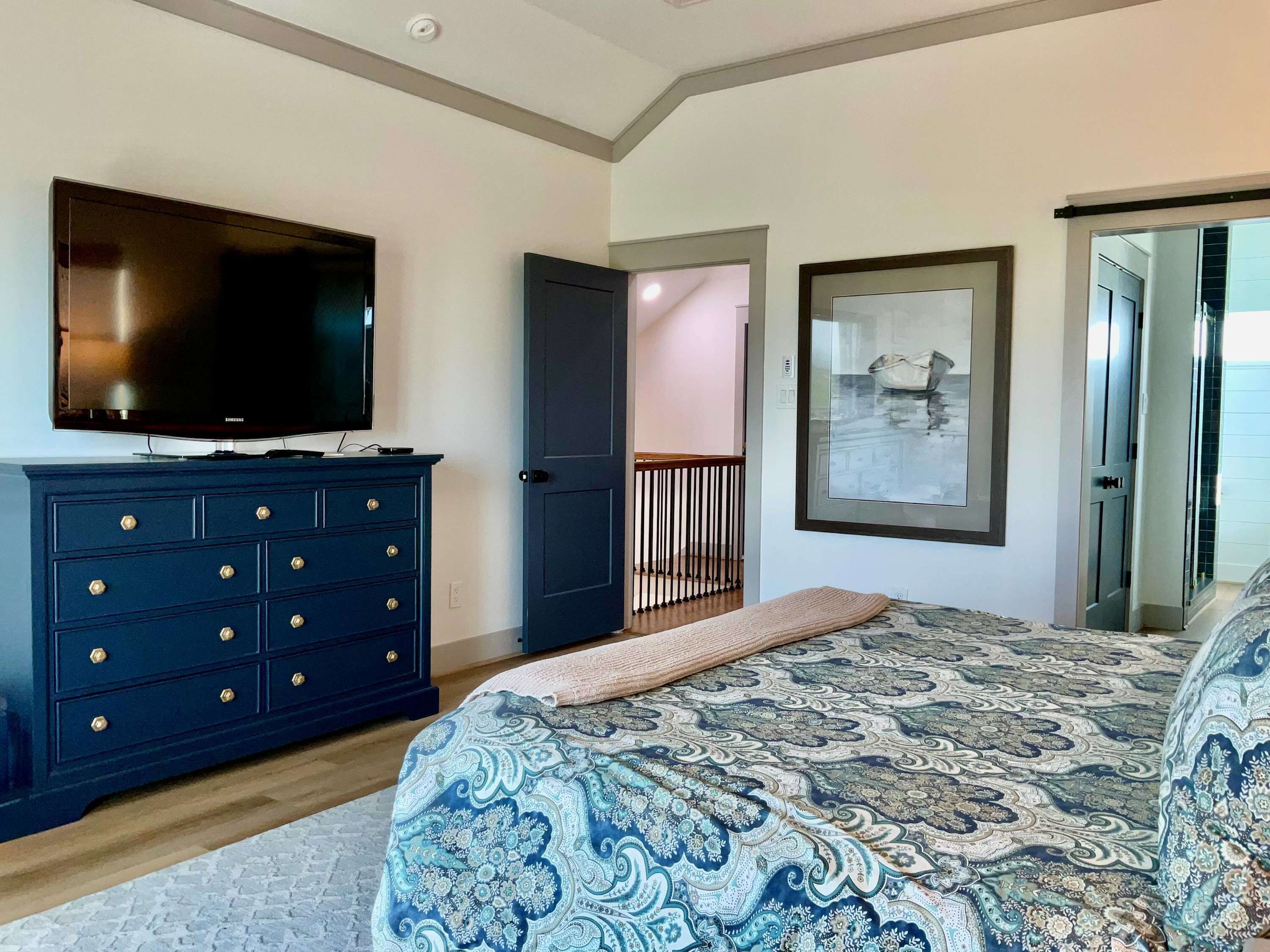 Een slaapkamer met een bed, een dressoir, een televisie, een lijst met een afbeelding en een deur naar een andere kamer.