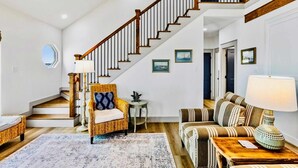 A spacious living area with a staircase, a wicker chair, a striped sofa, and a lamp. - Spoonbill at West Bay (Galveston)