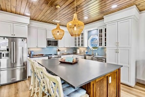 A modern kitchen with white cabinetry, a large island, hanging wicker pendant lights, and a stainless steel refrigerator. - Spoonbill at West Bay (Galveston)
