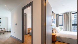 Ein modernes Hotelzimmer mit Holzboden, einem Bett mit weißen Leinen, einem Nachttisch und Blick auf die Stadt durch große Fenster.