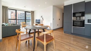Eine moderne Küche mit integrierten Geräten, ein Essbereich mit rundem Tisch und Holzstühlen sowie eine Sitzecke mit Blick auf die Stadt.