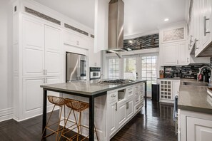A modern kitchen with white cabinetry, a stainless steel refrigerator, a gas stove, and a large island with two wicker stools.