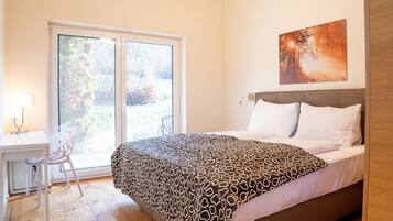 A bedroom with a bed, a desk, and a chair, a large window, and a painting on the wall.