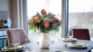 A dining table set with plates, glasses, and cutlery, a vase with flowers, and a view through a window. - Apartment Near Graz With Sauna (Premstätten bei Vasoldsberg)