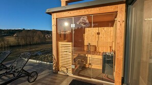 A wooden sauna with glass doors, a patio with metal chairs, and a view of a forested area. - Apartment Near Graz With Sauna (Premstätten bei Vasoldsberg)