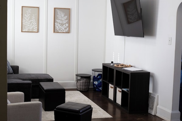 A modern living room with a sofa, ottomans, a TV mounted on the wall, and a small side table with decorative items. - Kenosha Retreat - Created to Help You Relax (Kenosha)